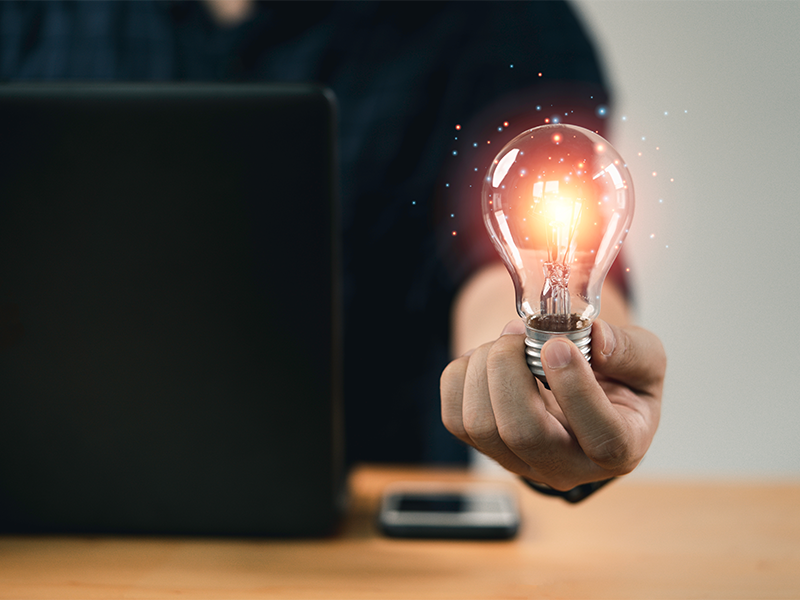 Man sitting in front of laptop holding a lighted bulb in an outstretched hand