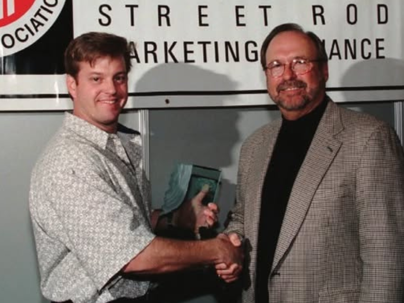 Two men smile and shake hands as they stand in front of a banner that reads “Street Rod Marketing Alliance.&quot;