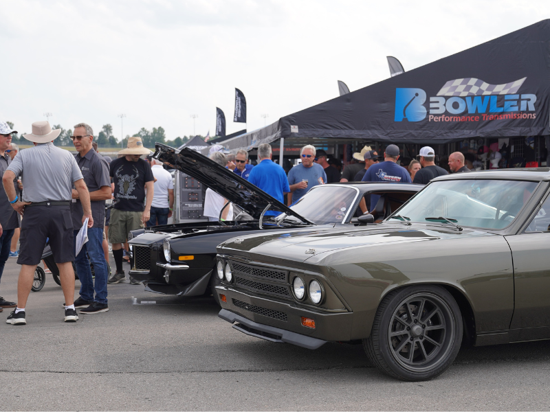 Crowd at Happy Hour at Bowler Transmissions during the Triple Crown of Rodding , with classic muscle cars on display—an olive-green coupe foreground, another with hood up—outside the Bowler tent.