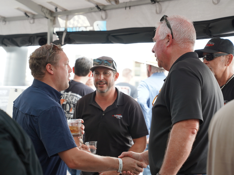 Triple Crown of Rodding networking scene under a vendor canopy as two attendees shake hands while others chat, one holding a drink.