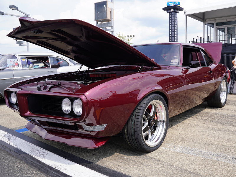 riple Crown of Rodding show car—deep burgundy classic muscle car with hood open and wide wheels—parked on the track for display.