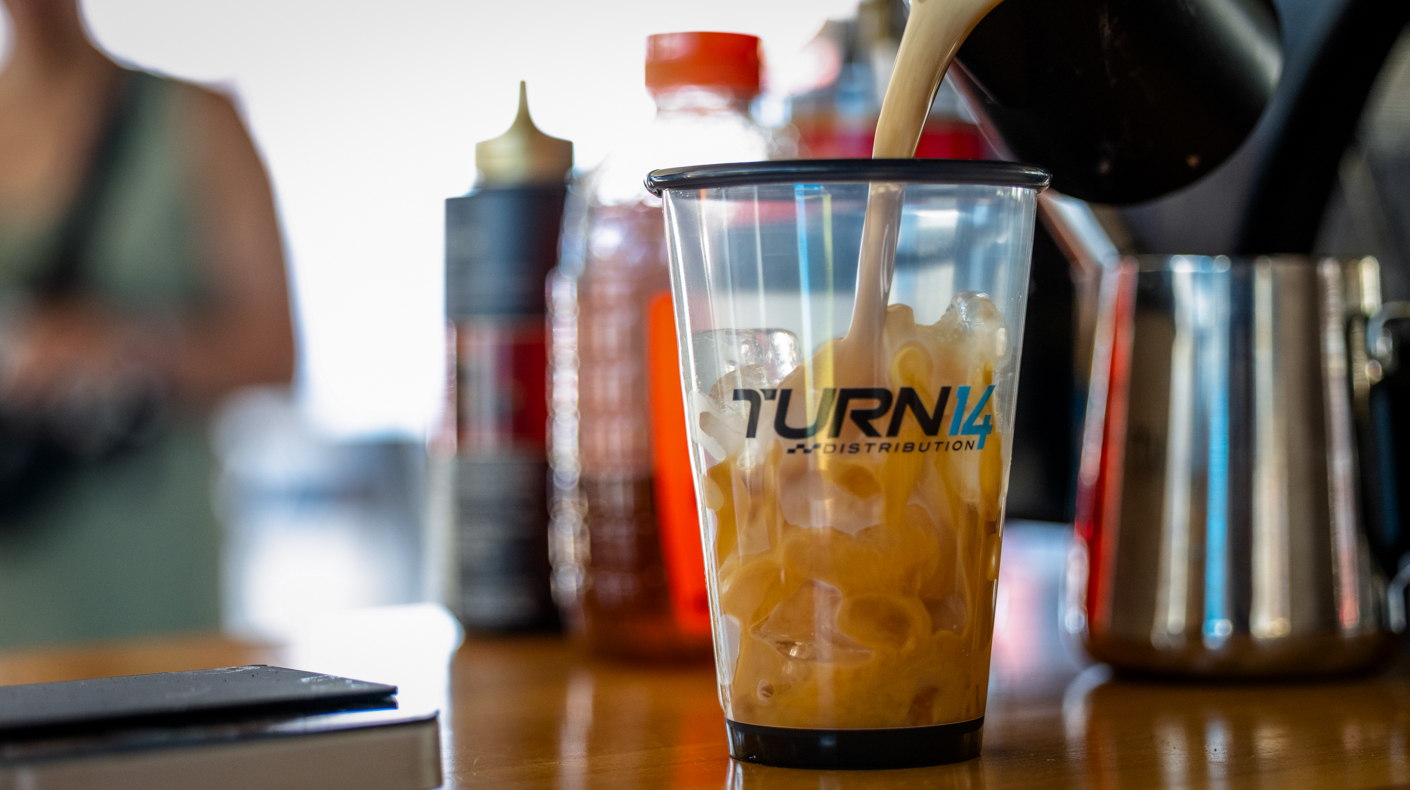 Coffee pouring into a cup of ice with the Turn 14 logo on it.